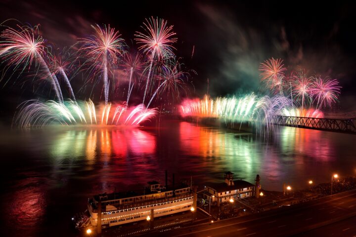 Colorful fireworks are displayed over a river at night, reflecting on the water; a docked riverboat and a bridge are visible in the foreground and background.