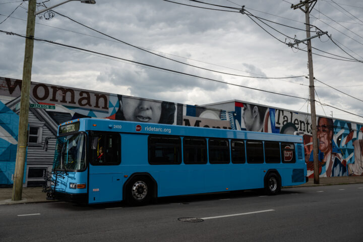 A blue city bus is parked on a street in front of a mural featuring faces and large text on a building under a cloudy sky.
