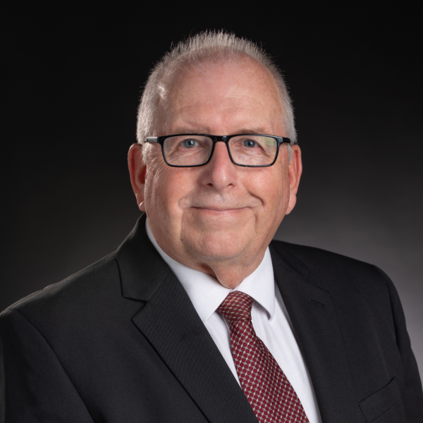 A middle-aged man with glasses in a black suit, white shirt, and red patterned tie poses against a dark background.