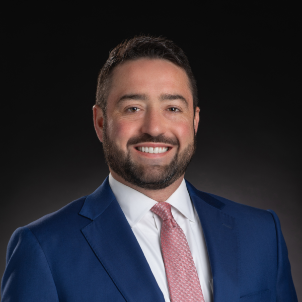 Man with dark hair and beard wearing a blue suit, white shirt, and pink tie, smiling in front of a plain dark background.
