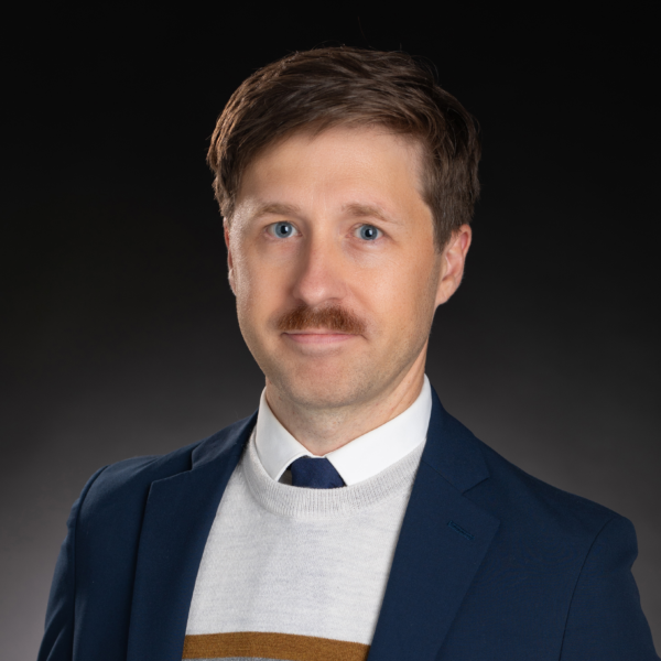 Man with light brown hair and mustache wearing a blue suit jacket, white shirt, tie, and sweater, posing against a dark background.