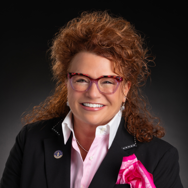 A woman with curly reddish hair, glasses, and a black blazer with a pink handkerchief smiles in front of a dark background.