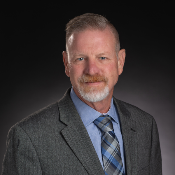 A middle-aged man with short gray hair and a beard, wearing a gray suit, blue shirt, and plaid tie, poses against a dark background.