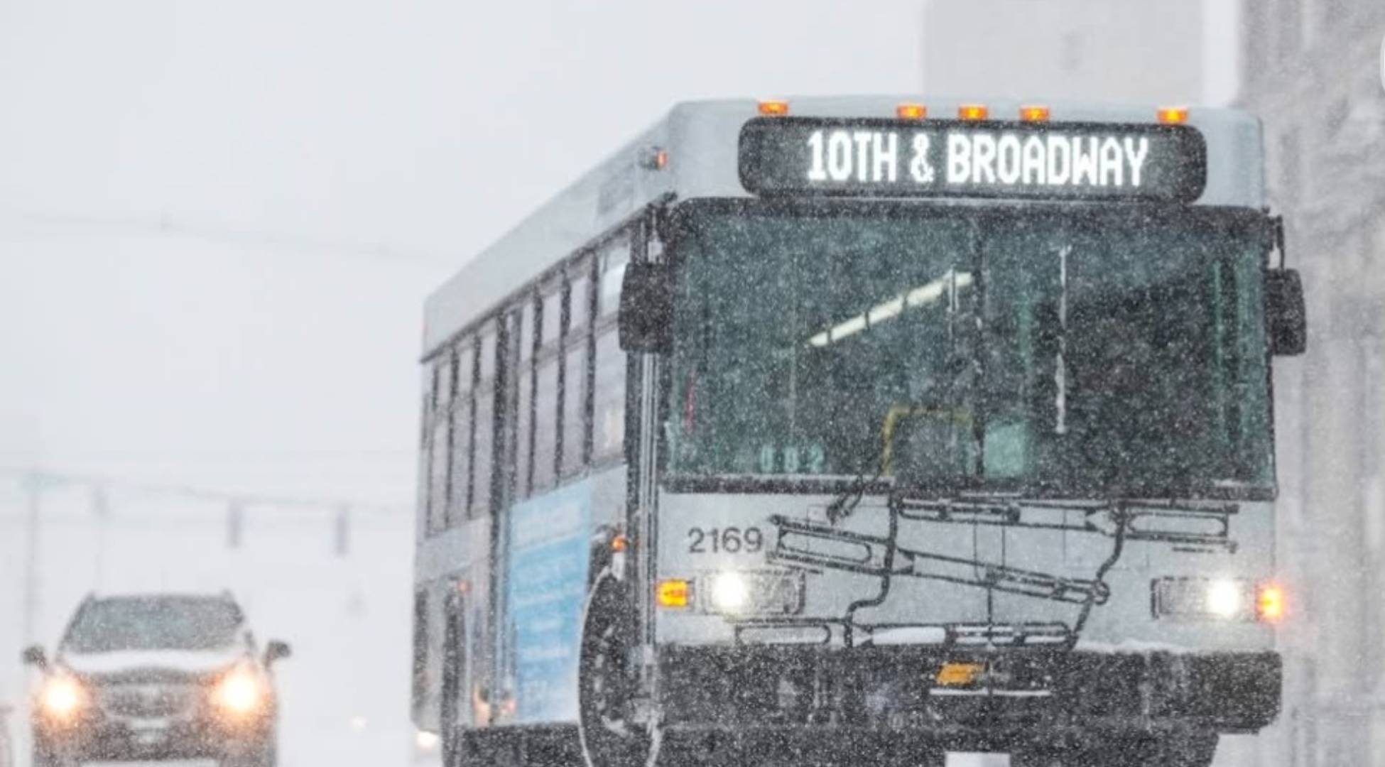 A city bus labeled "10th & Broadway" drives through heavy snow, with limited visibility. A car follows behind on the snow-covered street.