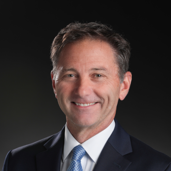 A middle-aged man in a dark suit, white shirt, and blue tie smiles at the camera against a dark background.