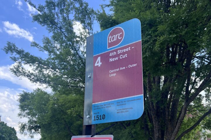 A TARC bus stop sign for Route 4 (4th Street - New Cut) stands on a sidewalk next to a street, with trees and parked cars in the background.