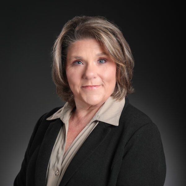 A woman with short, wavy brown hair wearing a beige blouse and black blazer poses for a professional headshot against a dark background.