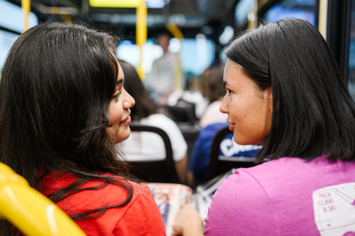 Two women sit side by side on a bus, facing each other and talking. Other passengers are visible in the background.