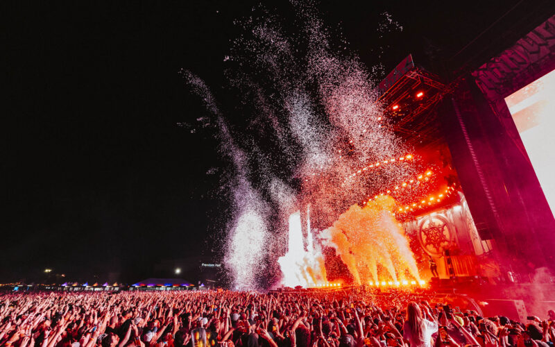 A large crowd at an outdoor concert at night with bright red stage lights, smoke, and confetti filling the air.