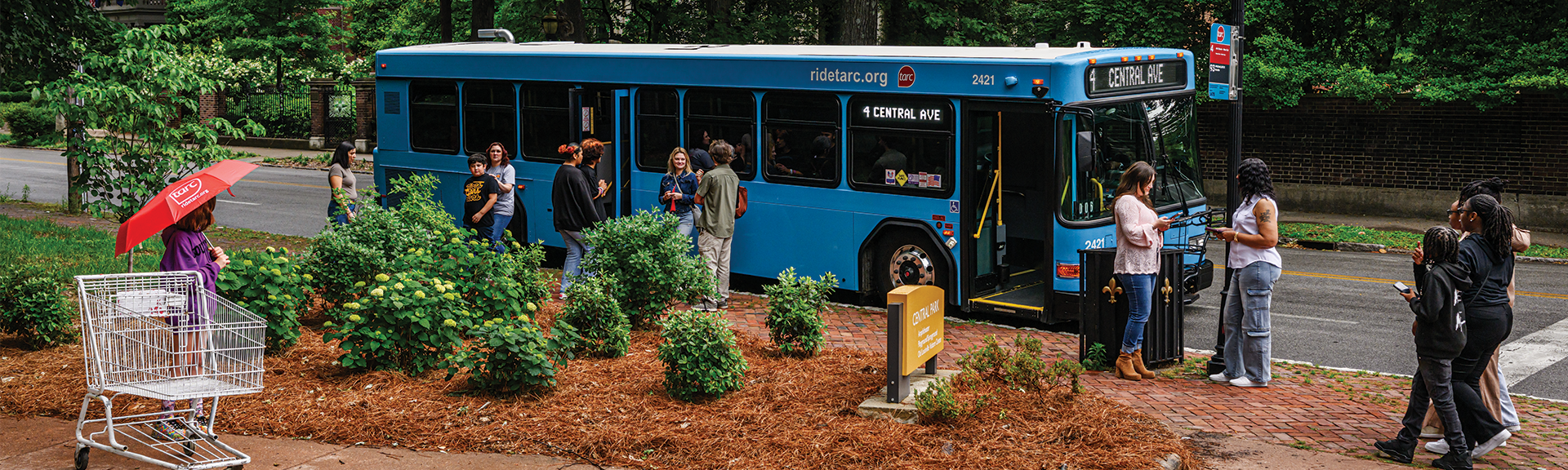 People wait at a bus stop as a blue city bus arrives. Some are boarding while others stand nearby. A red umbrella and an empty shopping cart are visible on the left side of the image.