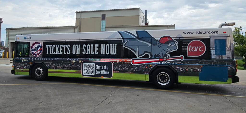 Side view of a city bus with an ad showing a flying bat in a baseball cap, promoting tickets for the Louisville Slugger Field, with QR code and website link.