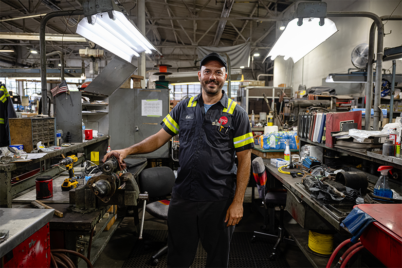 A mechanic in a uniform stands smiling in a cluttered auto repair shop with tools, parts, and workbenches visible around him.