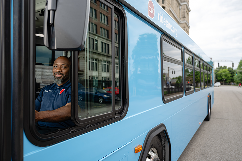 A bus driver in a blue uniform sits at the wheel of a blue city bus, looking out the open window on a city street during the day.