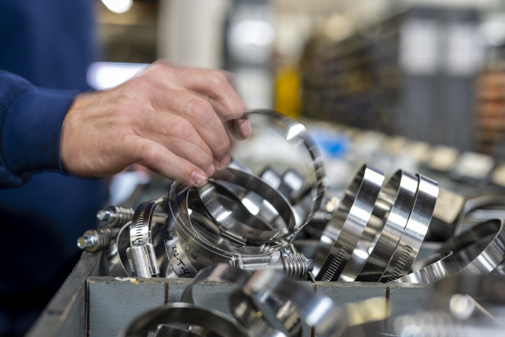 A person’s hand selects a metal hose clamp from a drawer filled with various metal clamps in a workshop or industrial setting.